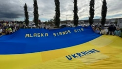 Anchoragede insanlar Ukrayinağa qoltutmaq içün büyük Ukrayina bayrağını tuta, Alâska ştatı, 2025 senesi avgustnıñ 14-ü, ABD ve Rusiye prezidentleriniñ körüşüvi arfesinde 