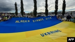 Anchoragede insanlar Ukrayinağa qoltutmaq içün büyük Ukrayina bayrağını tuta, Alâska ştatı, 2025 senesi avgustnıñ 14-ü, ABD ve Rusiye prezidentleriniñ körüşüvi arfesinde 