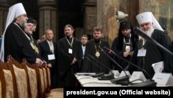 На цій світлині двоє ієрархів УПЦ (МП) під час Об'єднавчого собору: митрополит Вінницький і Барський Симеон (крайній зліва) і митрополит Переяслав-Хмельницький і Вишневський Олександр (в миру Олександр Драбинко, посередині на краю стола, об який спирається руками). Київ, 15 грудня 2018 року